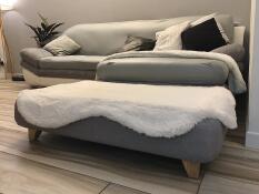 En grå säng med ett vitt fårskinnsöverdrag och fyrkantiga träfötter
